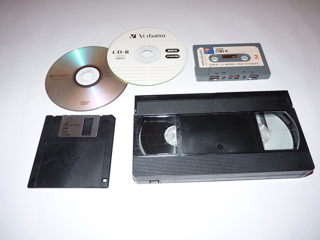 An image of obsolete media platforms: DVD, CD-R, cassette tape, VHS, and floppy disc.