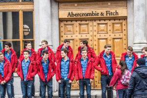 Two lines of 13 white male models, wearing the same outfit: jeans, a blue button down shirt, and a red sweater. They are standing in front of the doors of an Abercrombie and Fitch store.