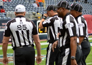 Four referees walking across a football field. They are wearing black and white striped shirts and hats.