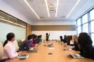 A group of people in a conference room turn their attention to a woman presenting.