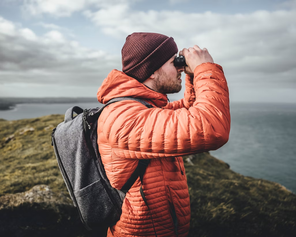 Man wearing a hat, orange jacket, and backpack looking out to the sea using binoculars.